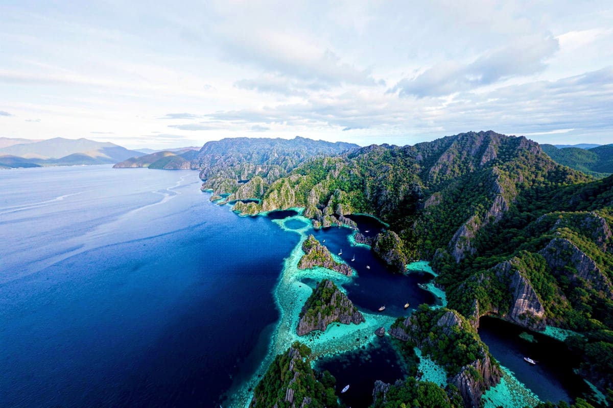 Coron Ultimate Island Day Tour Aerial view of Coron's limestone cliffs, turquoise lagoons, and forested islands with boats in crystal-clear tropical waters