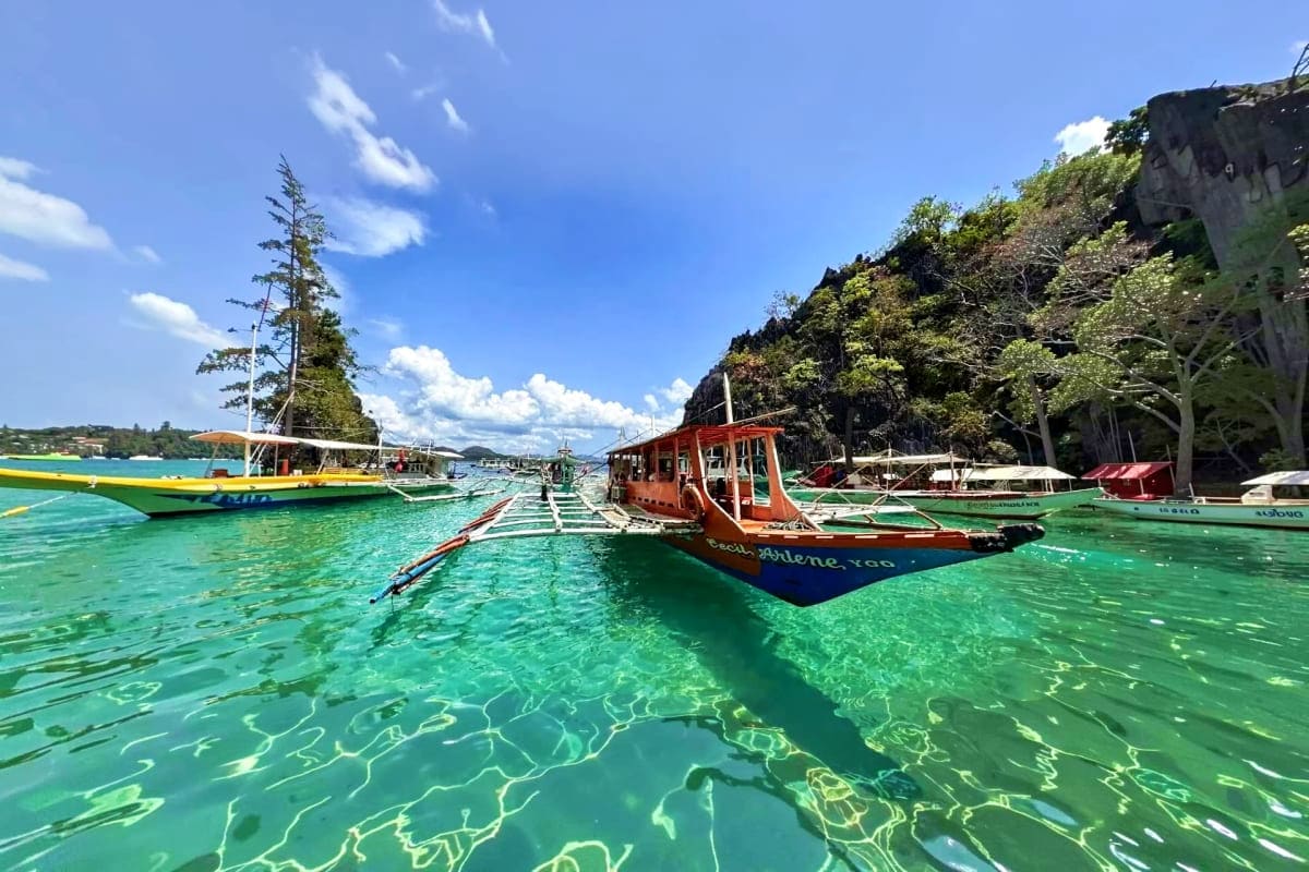 Coron Ultimate Island Day Tour Colorful traditional boats anchored in crystal clear turquoise waters near rocky limestone cliffs in Coron, Philippines