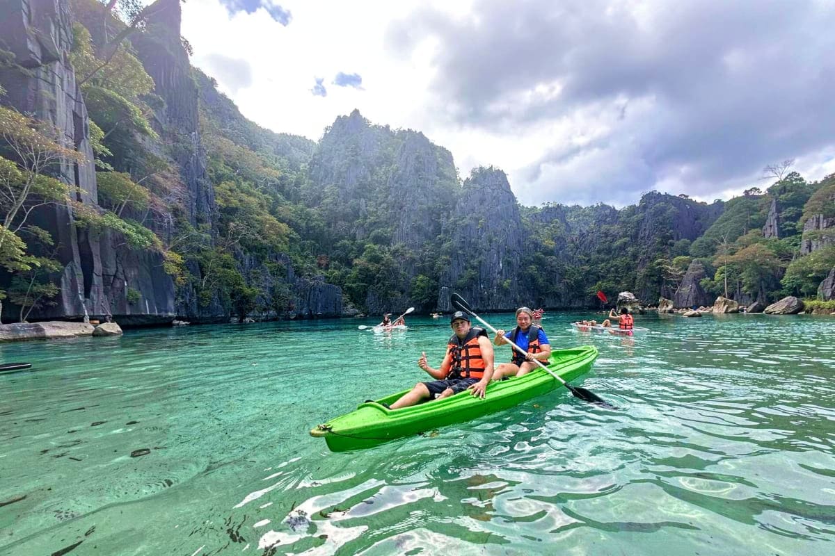 Coron Ultimate Island Day Tour Two tourists kayaking in a bright green kayak on turquoise water surrounded by towering limestone cliffs in Coron, Palawan