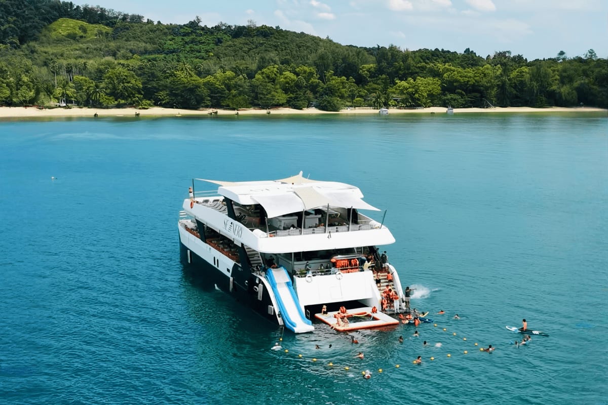 Luxury white and black catamaran with water slide anchored in turquoise bay, tourists swimming nearby, forested coastline in