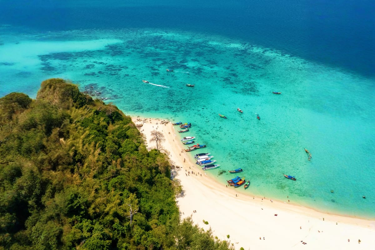 Aerial view of Phi Phi Island beach with colorful longtail boats, turquoise water, white sand, and lush green limestone cliff