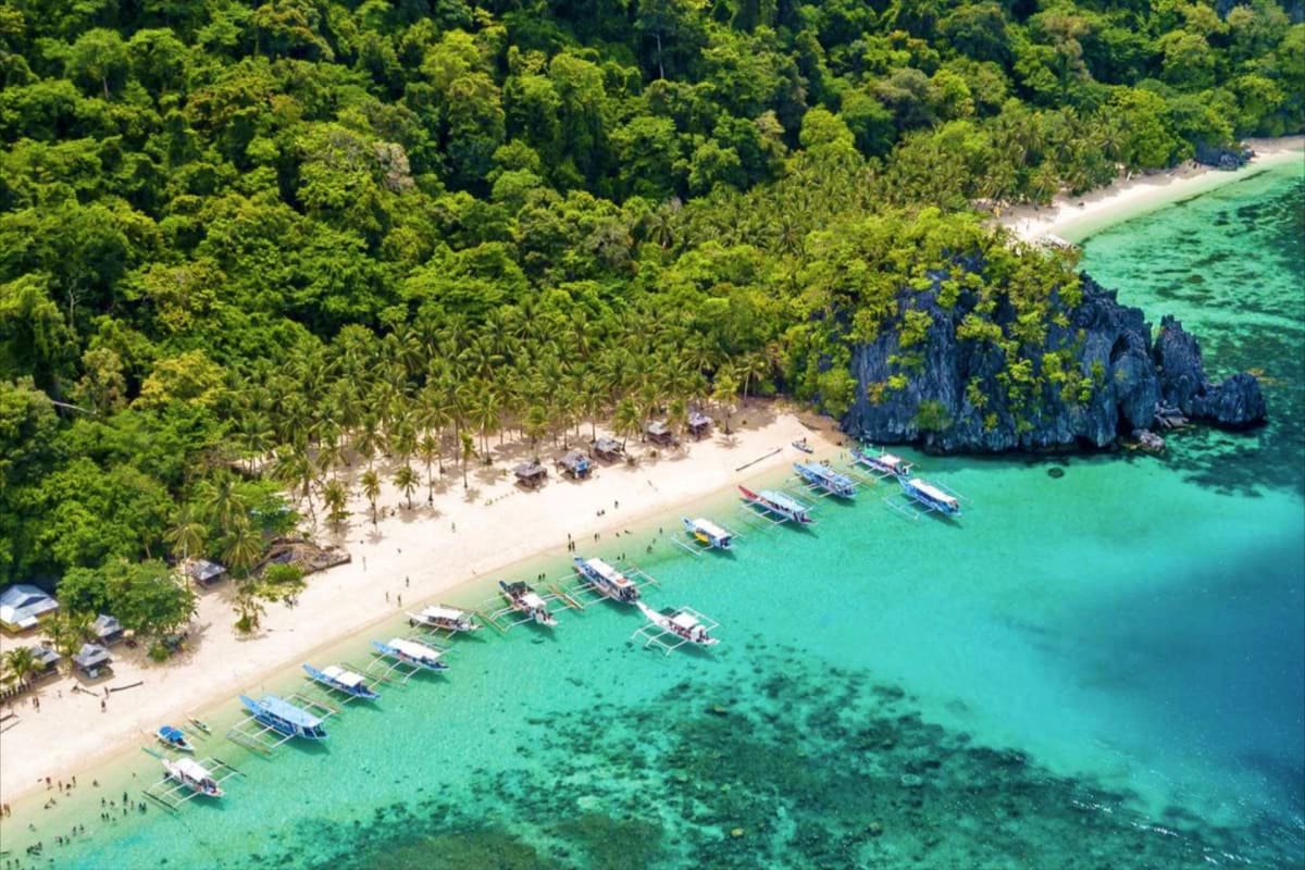El Nido Island Hopping Tour with Snorkeling Aerial view of tropical island beach with turquoise waters, moored boats, white sand, limestone cliffs, and dense green jungl