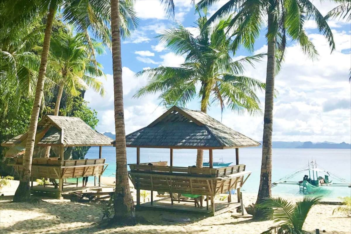 El Nido Tour D Lagoons and Beaches Tropical beach shelter with thatched roof, palm trees, wooden benches, and fishing boat in turquoise lagoon water