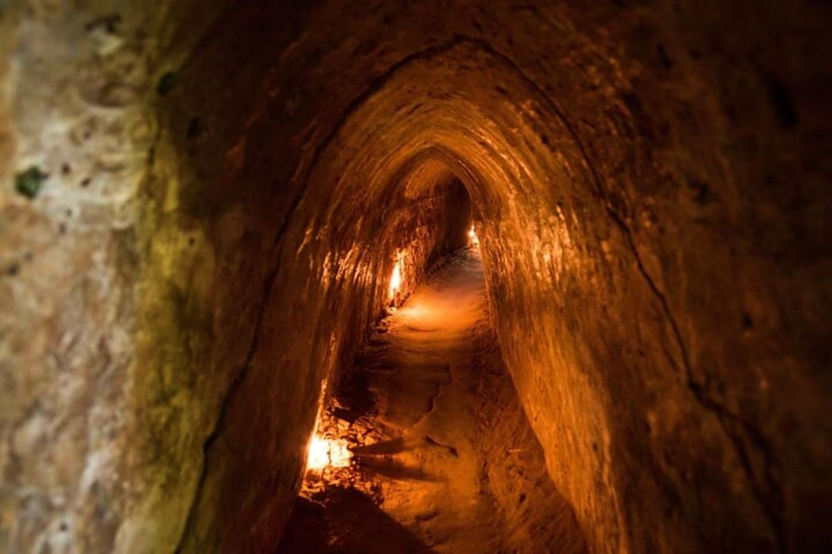 Cu Chi And Mekong Full Day Tour Narrow tunnel with brown clay walls illuminated by warm lighting, showing underground passage at Cu Chi Tunnels historic site