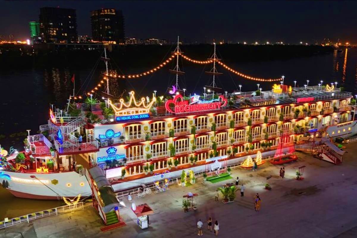 Illuminated riverboat with golden lights and "Ho Chi Minh" signage docked at night with passengers on pier