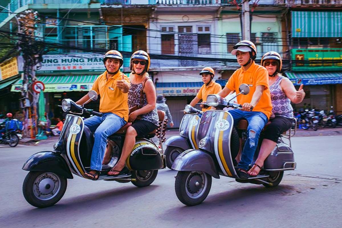 Saigon Street Food Tour By Motobike Tourists riding Vespa scooters on a Ho Chi Minh City street during a motorbike food tour, wearing yellow shirts and helmets w
