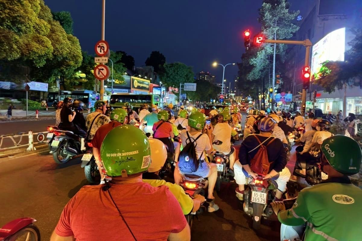 Saigon Street Food Tour By Motobike Large group of scooter riders wearing colorful helmets gathered on busy Ho Chi Minh City street at night for motorbike tour