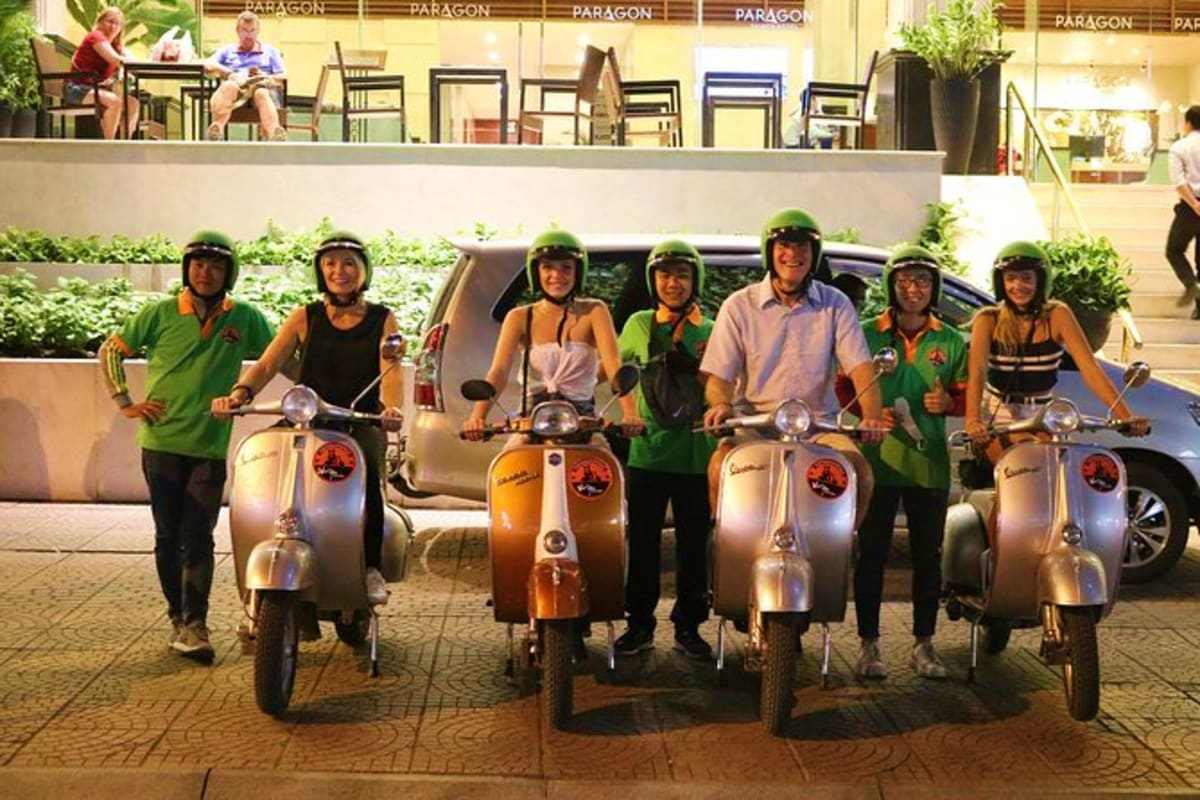 Group of seven people wearing green vests and helmets standing with vintage Vespa scooters in a urban plaza during daytime