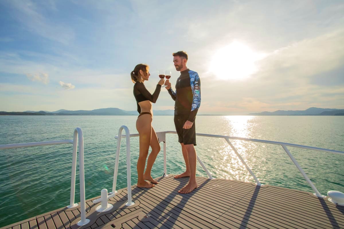 Couple toasting with drinks on a wooden pier with turquoise water and distant islands at sunset