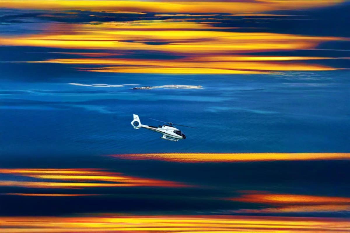 White helicopter flying over calm blue ocean waters at sunset with golden and orange sky reflections