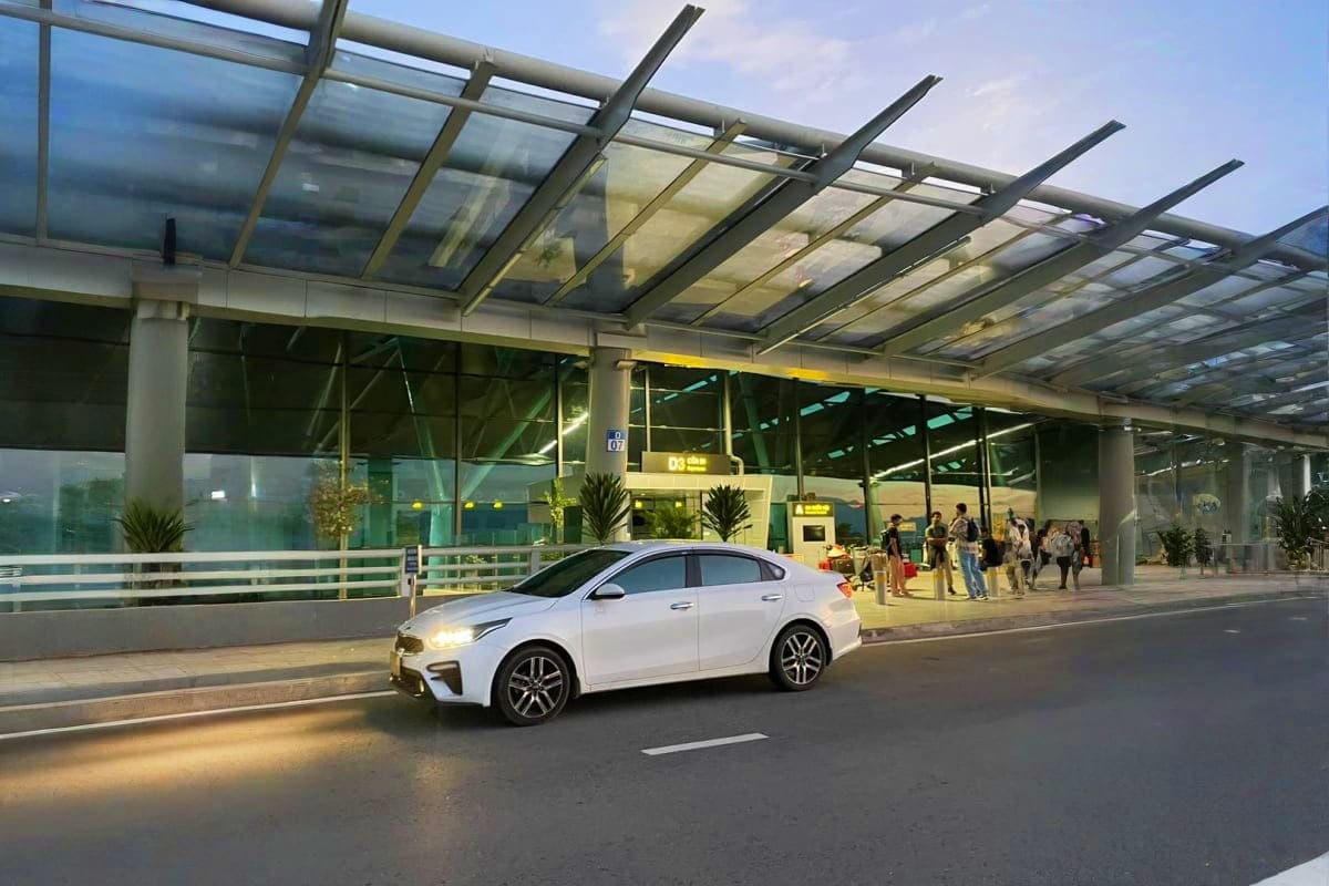 White sedan parked outside modern airport terminal with glass canopy and passengers waiting