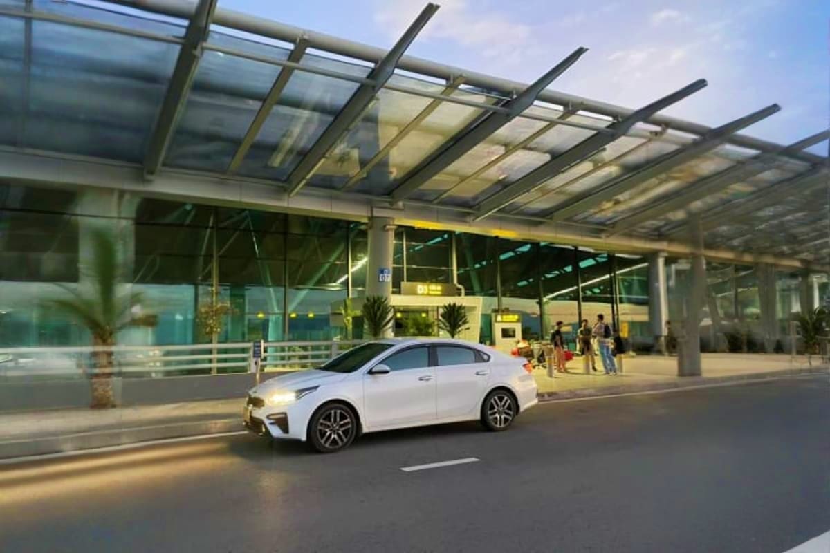 White sedan parked under modern airport terminal with glass canopy roof and passengers waiting nearby