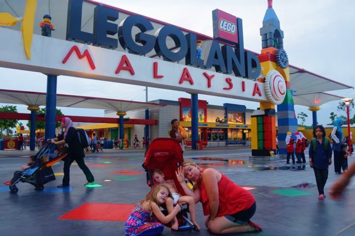 Family posing for photo at Legoland Malaysia entrance with iconic colorful LEGO brick tower and signage visible in background