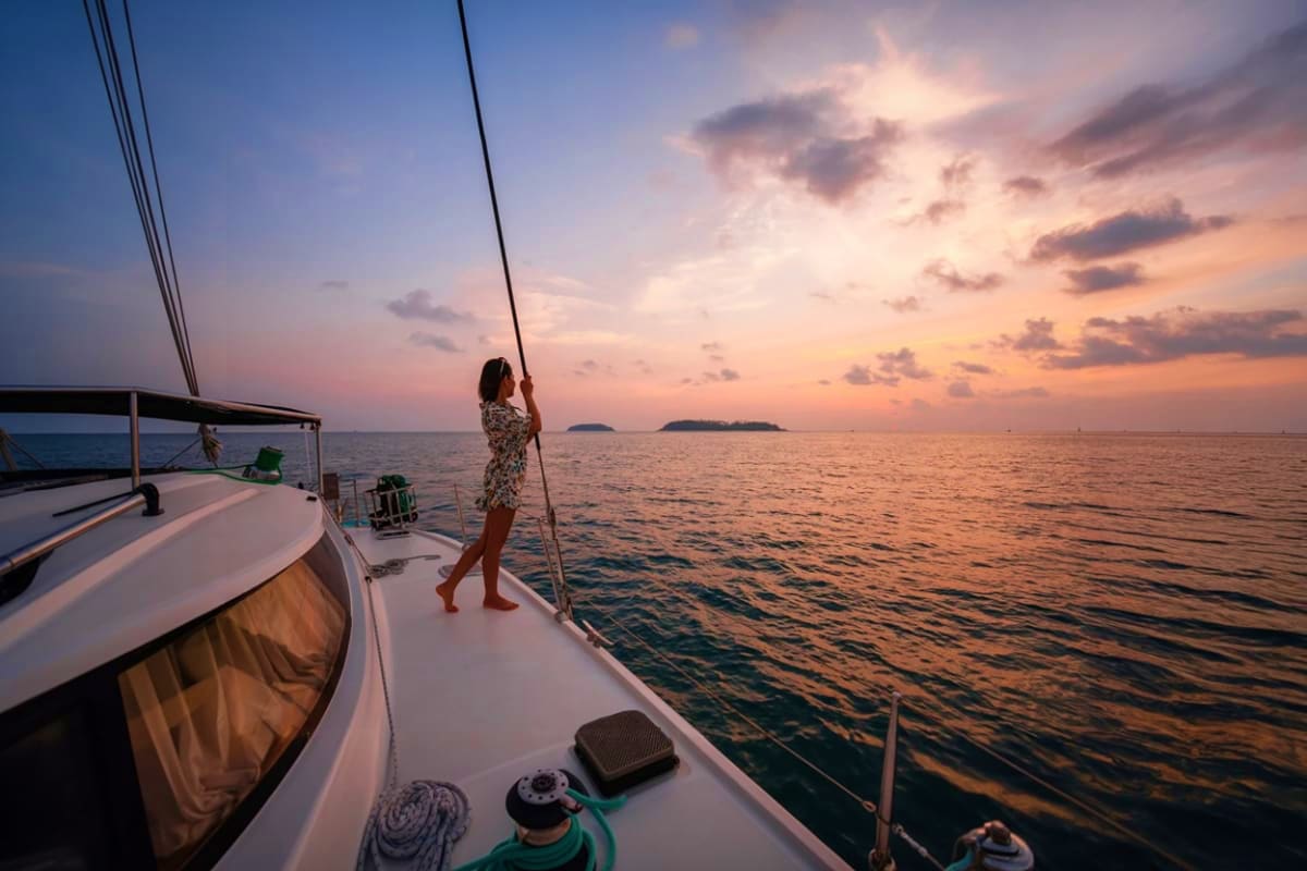 Woman holding sailboat rigging at sunset on catamaran with distant islands on horizon
