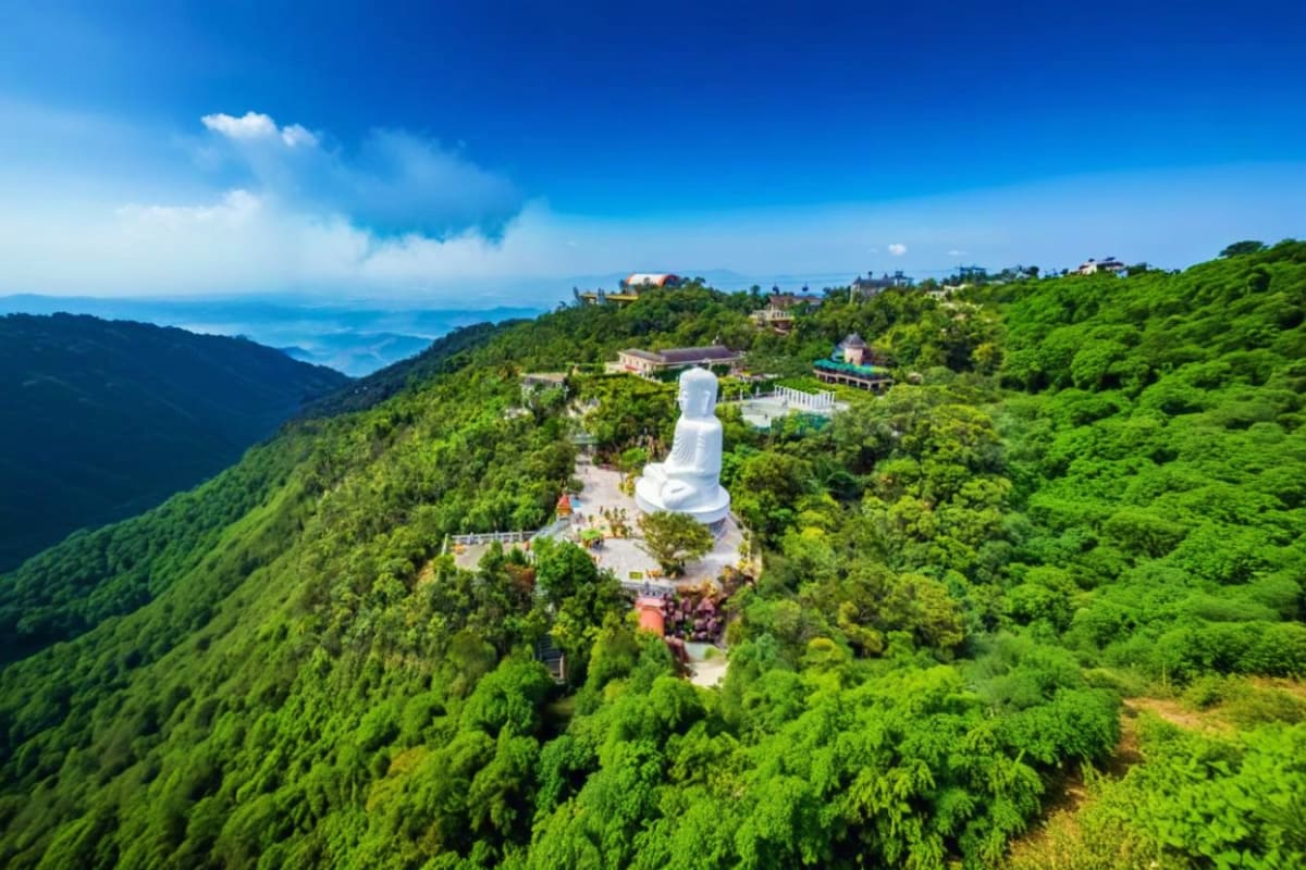 Danang and Hoi An City Tour Large white Buddha statue on mountaintop surrounded by lush green forests overlooking ocean and coastal landscape in Da Nang,