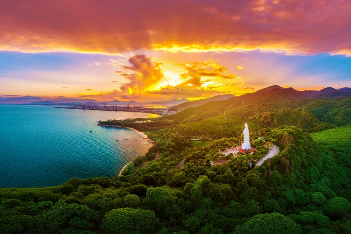 Aerial sunset view of white monument in forested hills overlooking turquoise bay with coastal city and mountains in backgroun