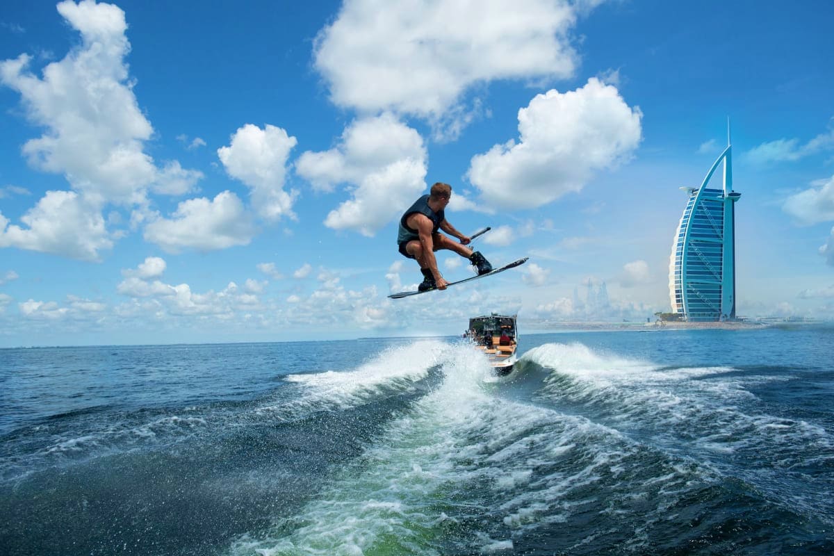Wake Boarding Experience 30 Min Wakeboarder airborne behind speedboat with Burj Al Arab sail-shaped hotel in background, Dubai coast with blue sky and white