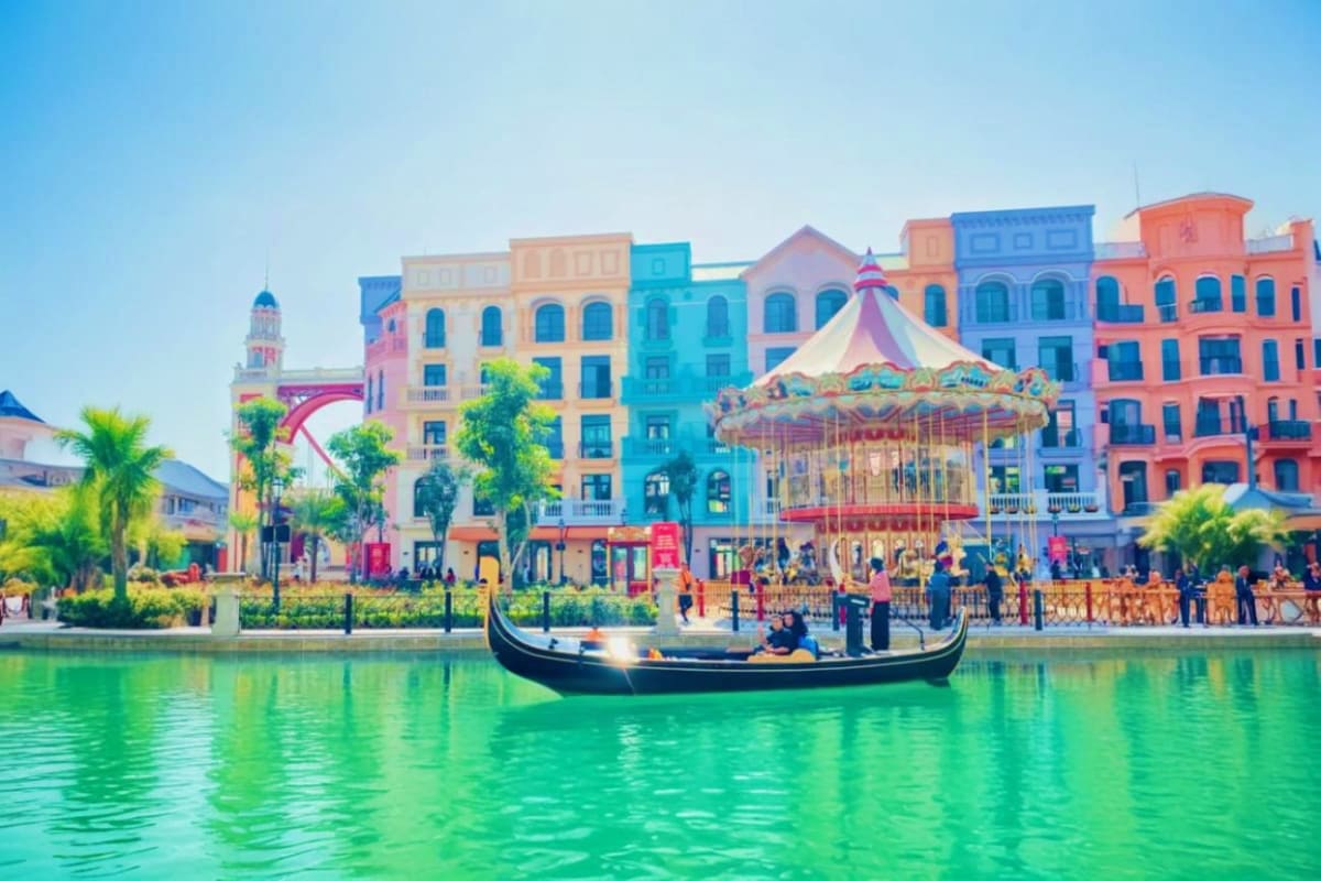 Gondola boat on turquoise water with colorful buildings, carousel, and palm trees in Venice-inspired theme park
