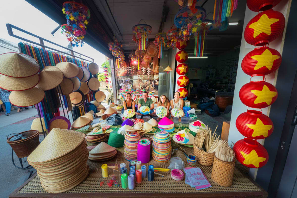Tourists sitting in a Vietnamese craft workshop surrounded by colorful conical hats, paper lanterns, and craft supplies durin