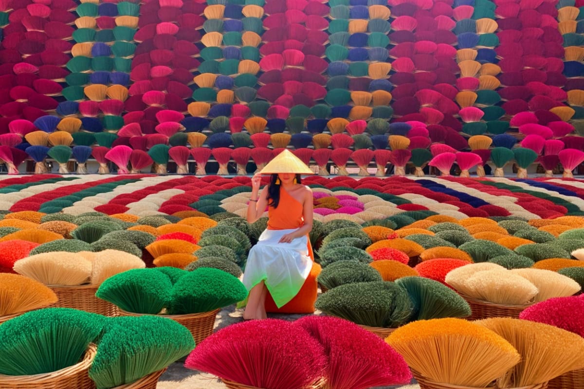Woman in orange shirt and conical hat sitting among colorful drying incense sticks and bamboo baskets at Hanoi incense villag