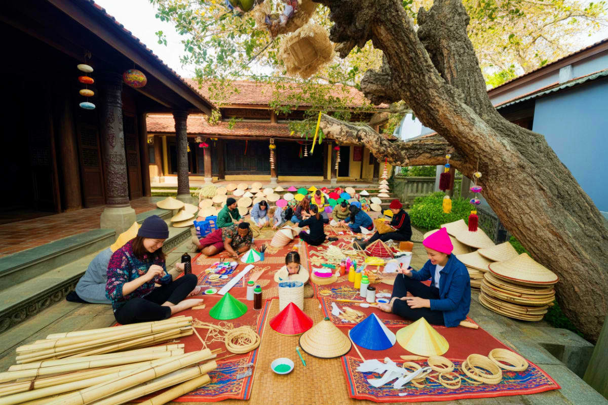 Group of visitors making colorful conical hats in courtyard of traditional Vietnamese village with ancient tree and wooden bu
