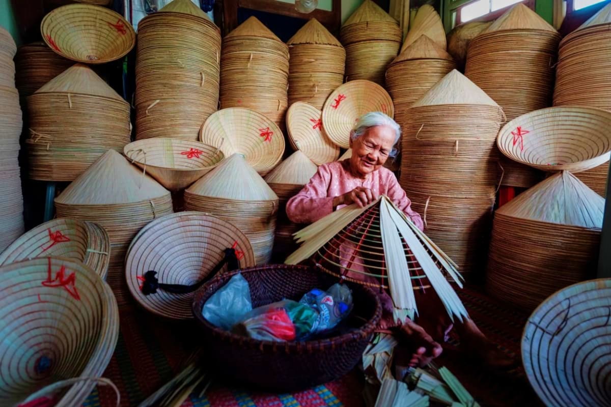 Elderly Vietnamese craftsman making traditional conical hats surrounded by stacks of woven palm leaf hats in Hanoi workshop