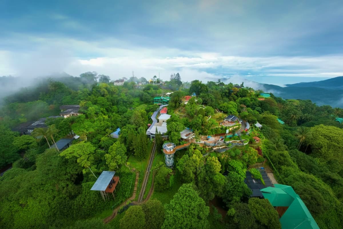 Aerial view of Penang Hill with lush green forest, winding train tracks, colorful roofs, and mountains in the background unde
