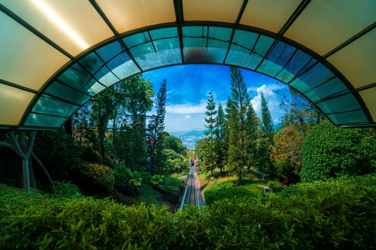 View through glass tunnel of Penang Hill Train tracks winding through lush tropical forest with blue sky and distant valley s