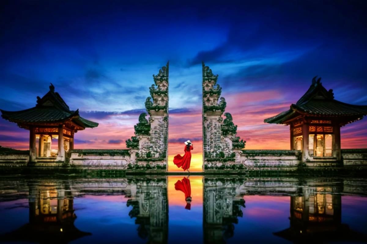 South Bali Tour Woman in red dress standing between ornate stone gates at sunset, reflected in still water at Bali temple entrance