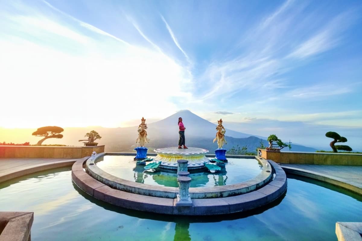 South Bali Tour Woman standing on circular fountain platform with Mount Agung volcano in background and manicured gardens at Bali temple sanc
