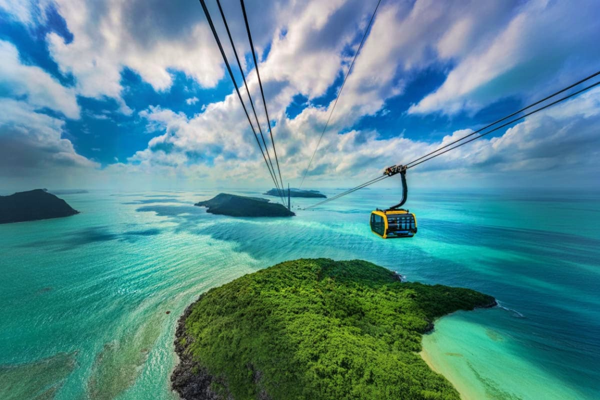 Cable car with yellow cabin suspended over turquoise sea and lush green island with distant islands under blue cloudy sky