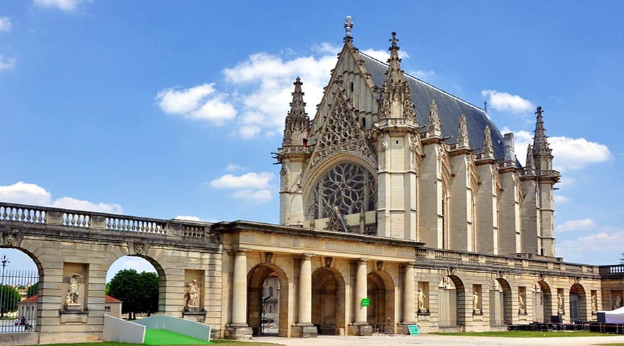 Gothic chapel with ornate stone facade, rose window, and arched colonnade entrance under blue sky