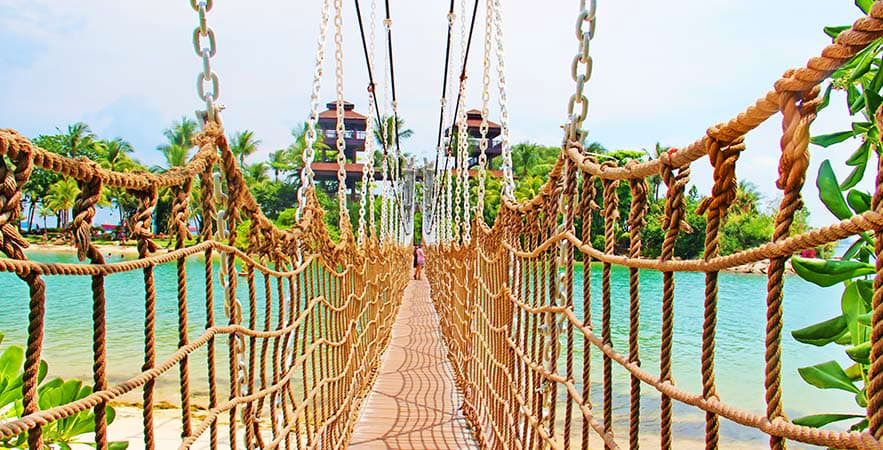Full Day Sentosa Island Tour Suspended rope bridge crossing turquoise water with tropical island vegetation and palm trees in background on Sentosa Island