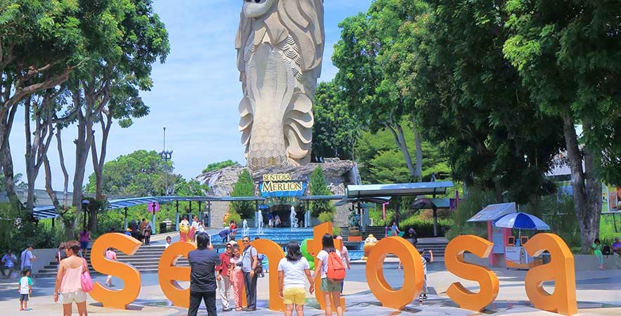 Visitors posing with large orange "Sentosa" letters at Merlion statue in Sentosa Island park with trees and blue structures i