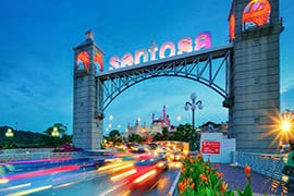Full Day Sentosa Island Tour Sentosa Gateway arch with illuminated sign at dusk, light trails from passing vehicles, and Singapore skyline in background