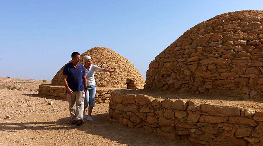Al Ain City Tour from Abu Dhabi Two tourists exploring ancient mud-brick beehive tombs at archaeological site in Al Ain desert landscape