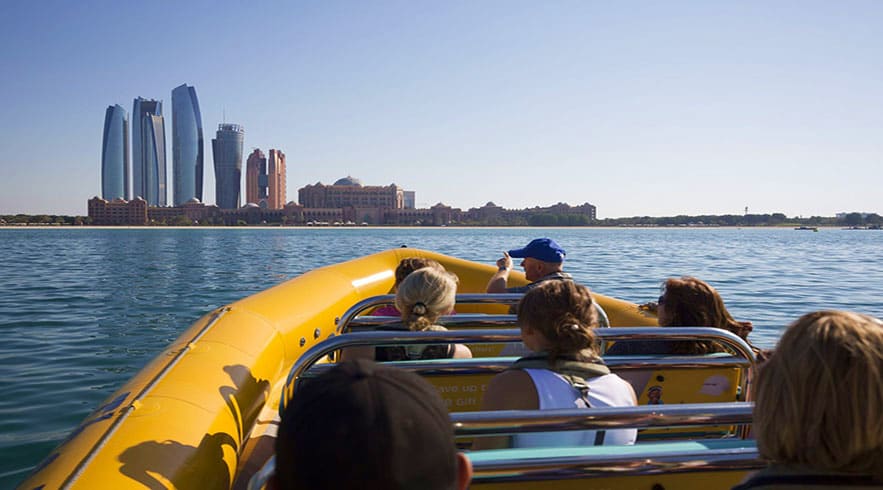 Tourists riding in a yellow speedboat with Abu Dhabi skyline and high-rise buildings visible across the blue water in the bac