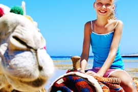Woman in blue swimsuit sitting on beach next to camel, holding colorful striped blanket, clear blue sky and sea in background