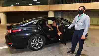 Man in business attire standing next to a black sedan with open door at indoor parking facility, Riyadh Airport