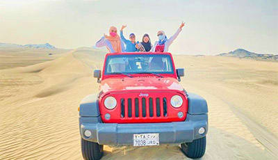 Group of tourists with arms raised on red Jeep driving through sandy desert landscape during dune bashing safari