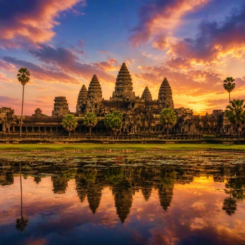 Angkor Wat temple complex reflected in water at sunset with palm trees and golden sky in Cambodia