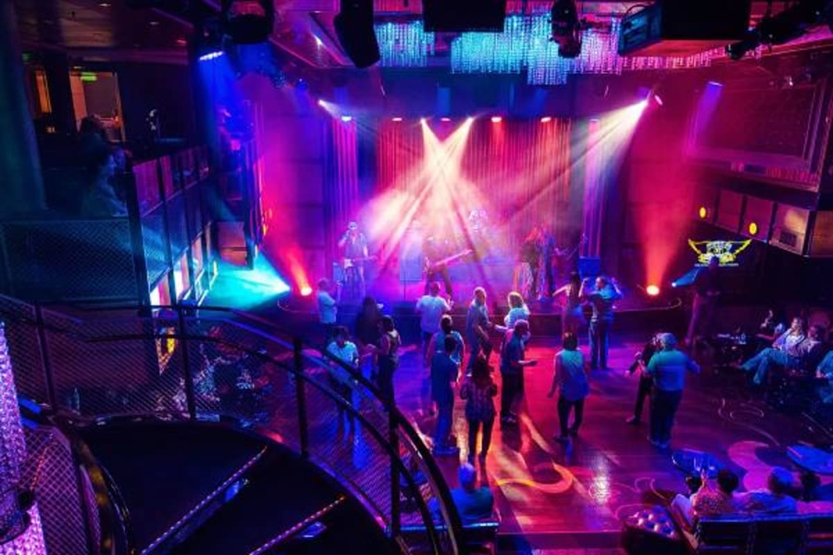 Concert venue on cruise ship with colorful stage lighting, performers, and guests dancing on multi-level dance floor