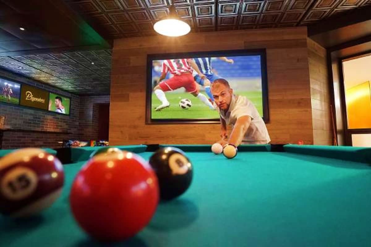 Man playing pool in cruise ship recreation room with billiard balls on teal table and soccer match displayed on screen