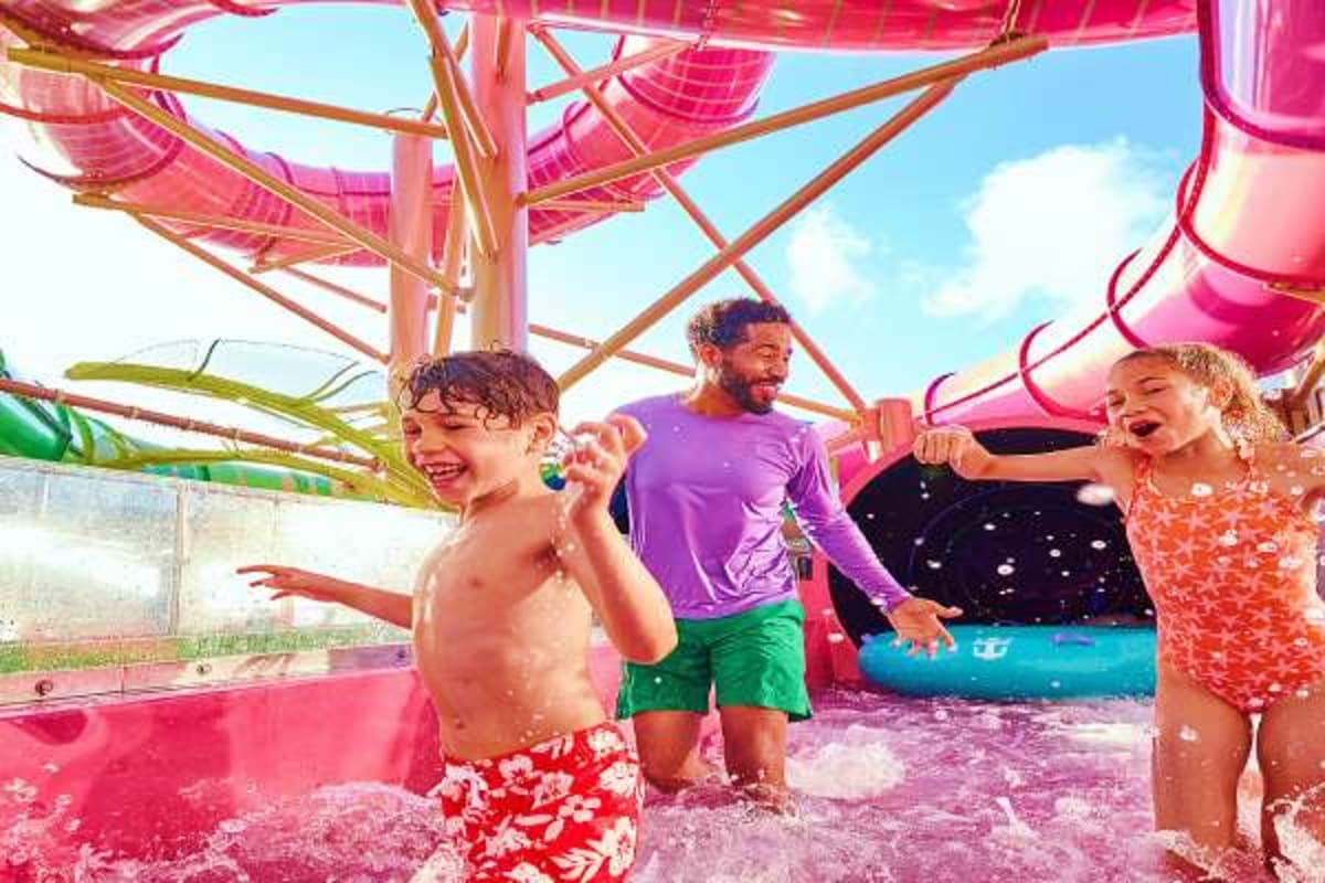 Family enjoying splash pad and water activities at outdoor pool area on cruise ship with pink canopy structures overhead