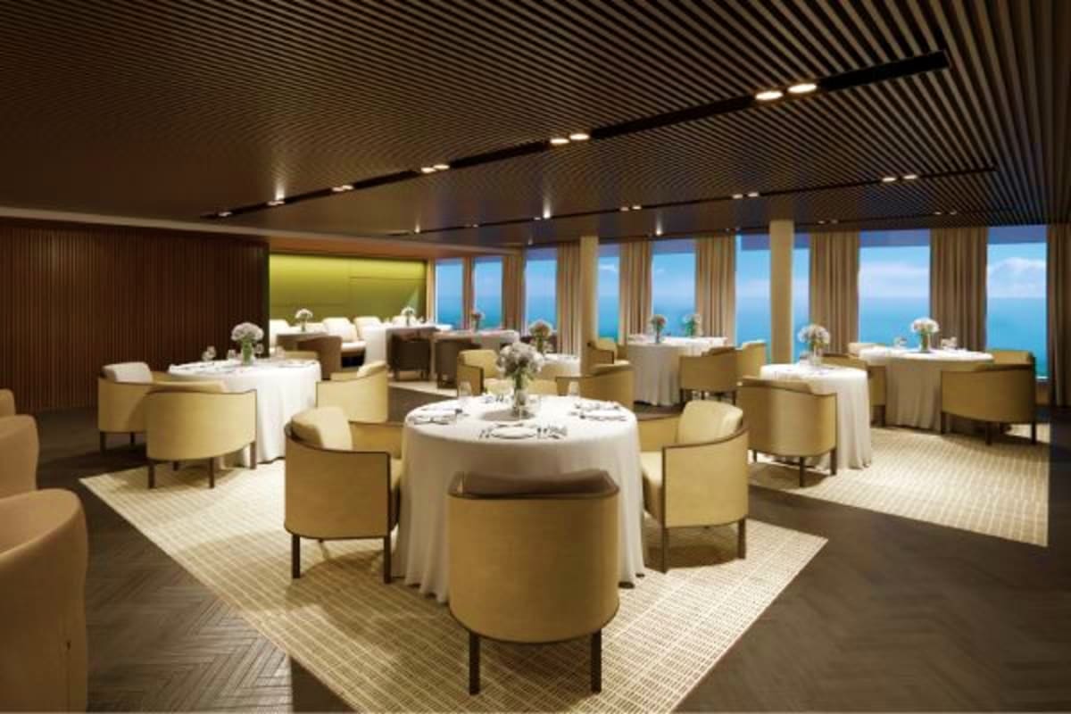 Luxurious cruise ship dining room with round tables, cream-colored chairs, floor-to-ceiling windows overlooking ocean and blu
