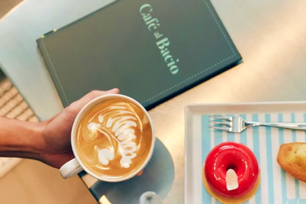 Person holding cappuccino with latte art at café table with cruise brochure and glazed donuts