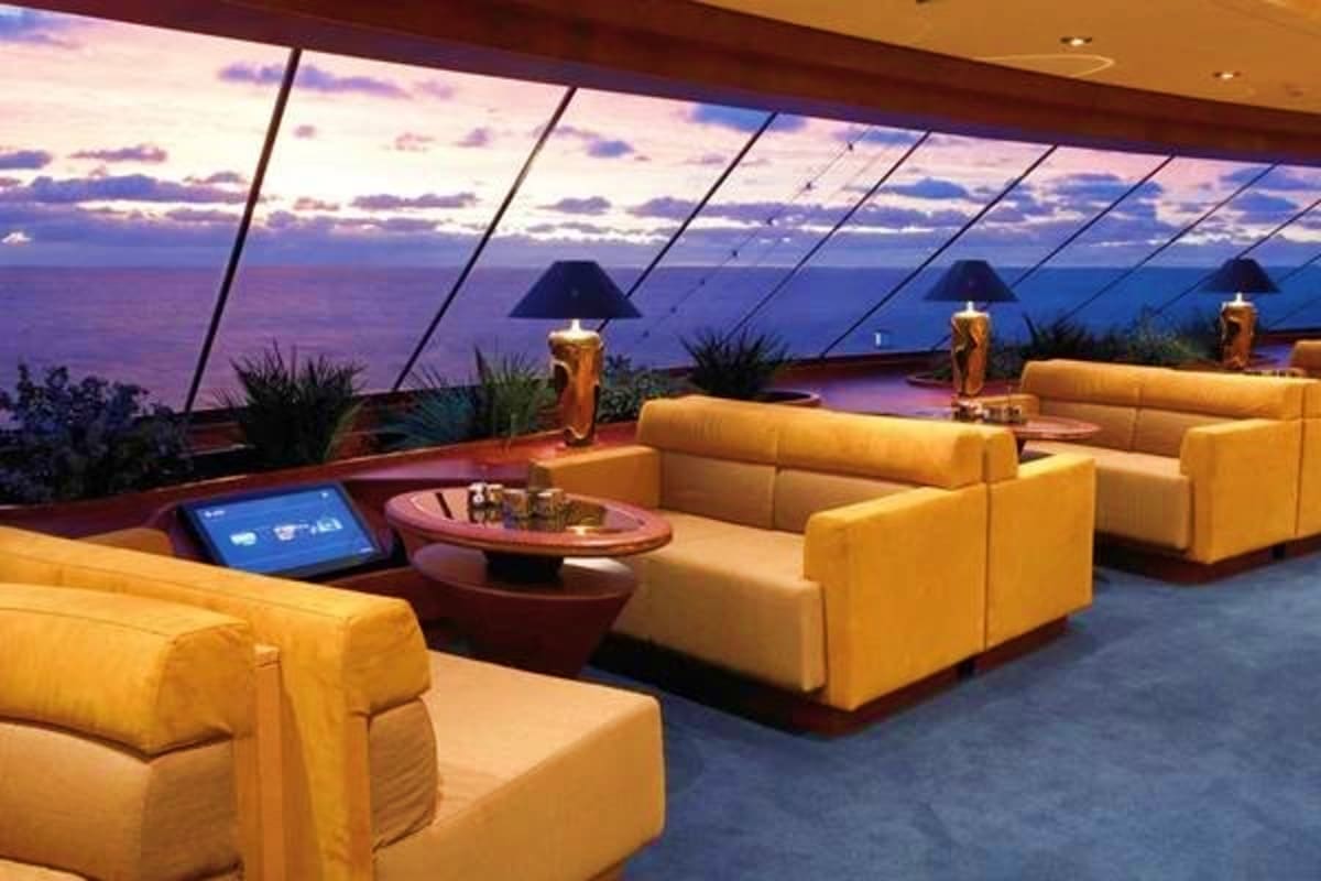 Modern cruise ship lounge with yellow seating, ocean and sunset view through floor-to-ceiling windows at dusk