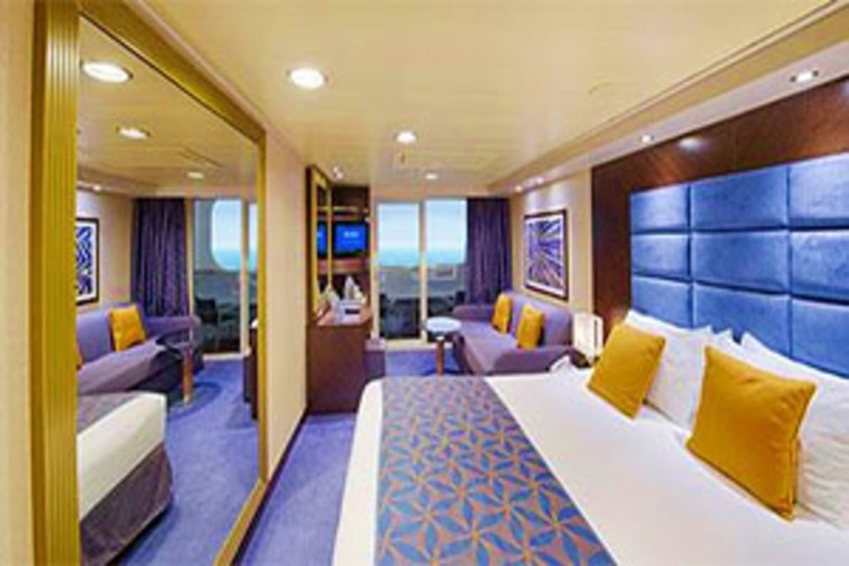 Luxury cruise ship stateroom with balcony view, double bed with gold pillows, seating area, and ocean horizon visible through