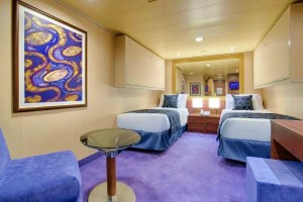 Spacious cruise ship cabin with two beds, purple carpet, blue and gold abstract artwork, side tables with lamps, and seating 