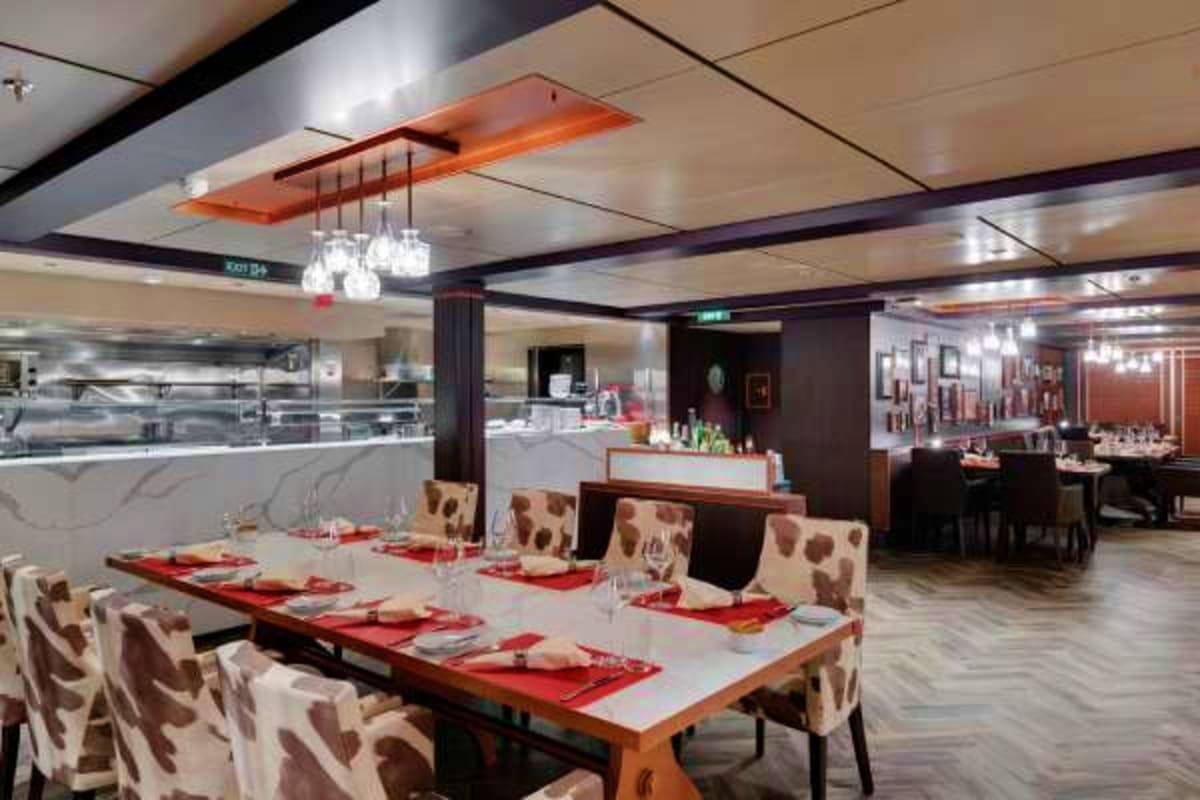 Modern cruise ship dining room with long table set with red napkins, patterned chairs, open kitchen view, and contemporary ce